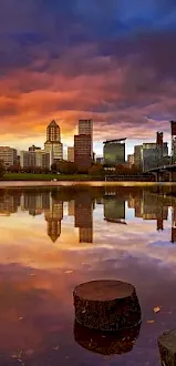 A dusk city skyline reflected in a calm river, with a bridge, train tracks, and rocky shoreline in the foreground.