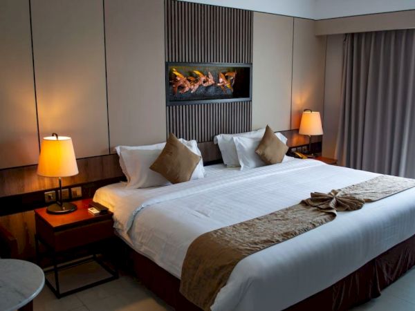 A modern hotel bedroom with a neatly made bed, bedside lamps, and an electric fireplace behind.