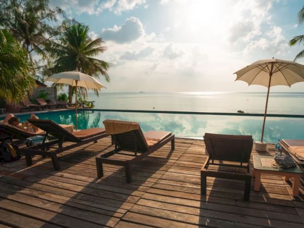 A tropical beachfront deck with sun loungers, umbrellas, and calm ocean under a partly cloudy sky.