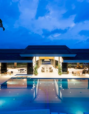 An illuminated outdoor hotel pool at dusk, framed by a modern building under a blue, cloud-filled sky.