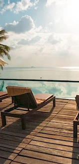 A beachside resort deck with lounge chairs and umbrellas overlooking calm ocean water under a bright, cloudy sky.