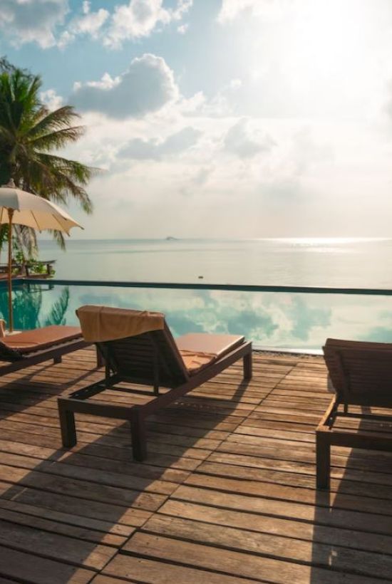 A tropical beach resort scene with lounge chairs, umbrellas, and a deck overlooking calm ocean waters under a cloudy sky.