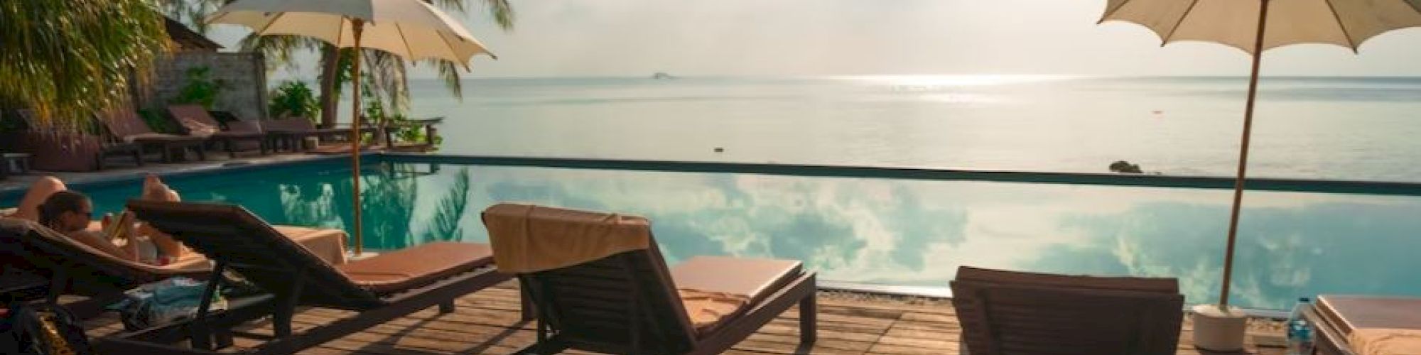 A tropical beach resort deck with lounge chairs under umbrellas, overlooking calm ocean waters at sunset, with palm trees framing the view.