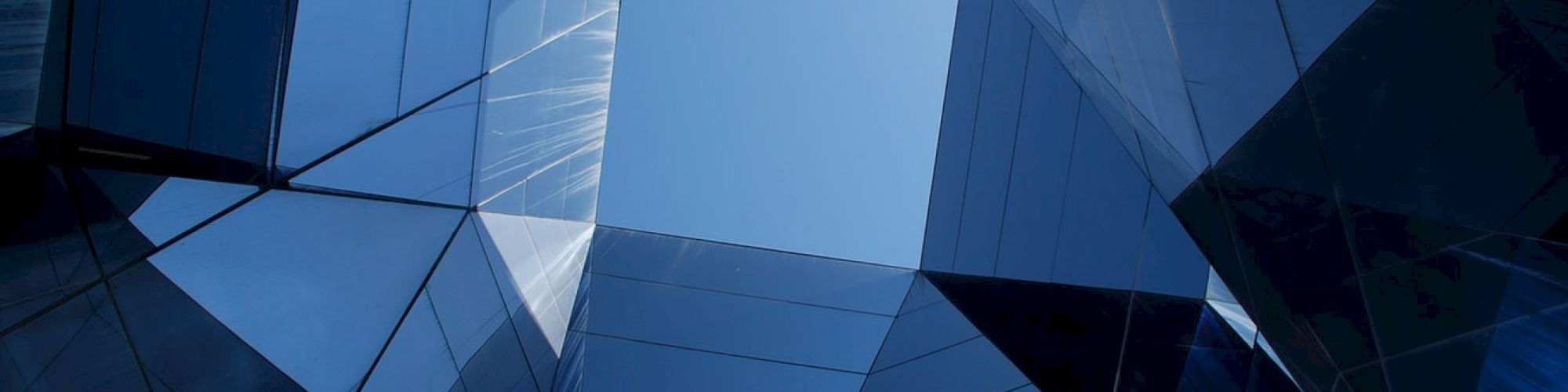 A close-up of a blue, faceted glass-and-metal skyscraper fa&ccedil;ade with sharp geometric reflections and panels.