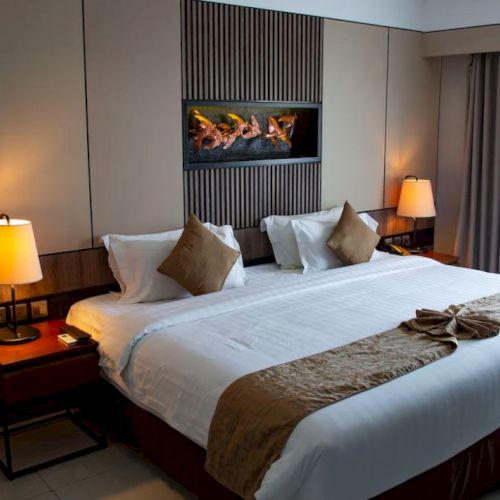 A cozy hotel room: neatly made bed with pillows, bedside lamps, and a wall fireplace glowing with flames.