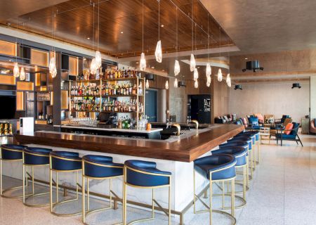 A modern hotel bar with a long wooden counter, backlit shelves of bottles, hanging pendant lights, and lounge seating.