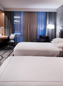A modern hotel room with two neatly made beds, a desk chair, TV, and curtains near a city-view window.