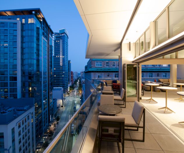 Modern cityscape at dusk from a high-rise terrace, showing outdoor lounge seating and glass doors overlooking streets and tall buildings.