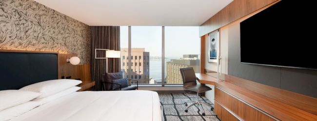 Modern hotel room with a large bed, wood paneling, a desk chair, and wall TVs, looking out over city buildings.