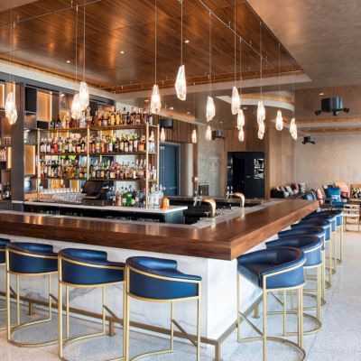 Modern hotel bar with a stocked back shelf, brass stools, pendant lights, and a lounge area with sofas in warm wood lighting.