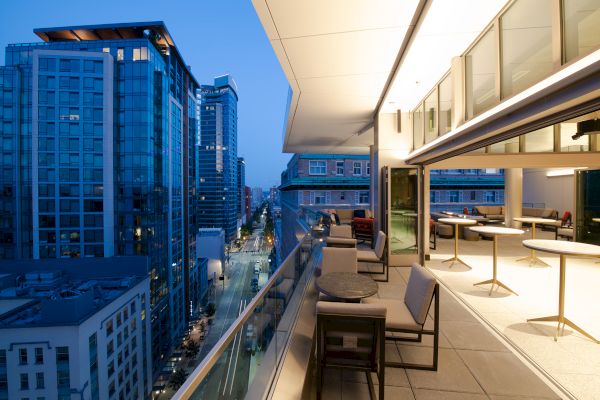 A modern rooftop terrace overlooks a downtown street, with lounge chairs and tables beside glass doors under a blue evening sky.