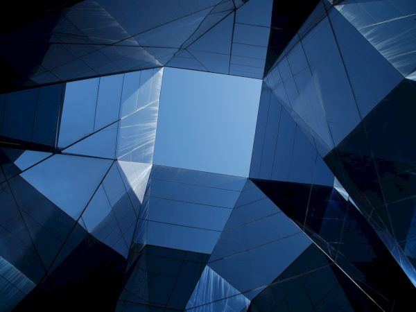 A futuristic building&rsquo;s blue glass facade forms sharp geometric triangles and polygons overhead.