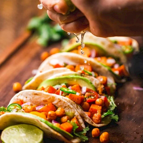 A hand pours sauce over crunchy tacos packed with veggies, beans, cilantro, and lime on a wooden table.