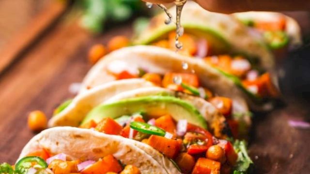 A hand pours sauce over crunchy tacos packed with veggies, beans, cilantro, and lime on a wooden table.