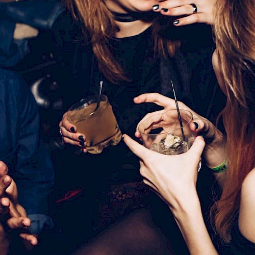 A dark party scene where people with face piercings sip drinks through straws from small cups, smiling and posing together.