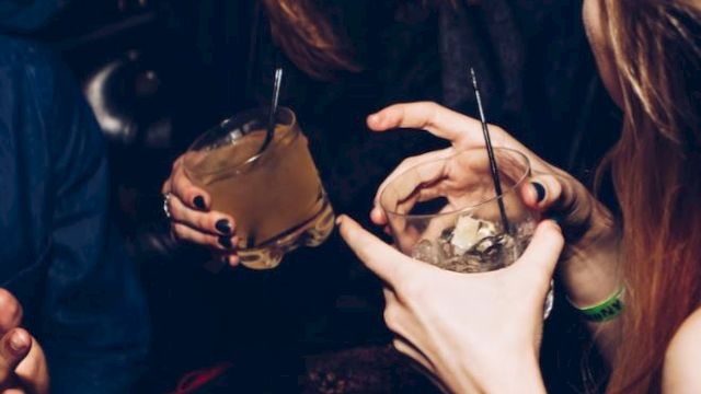 A dark party scene where people with face piercings sip drinks through straws from small cups, smiling and posing together.