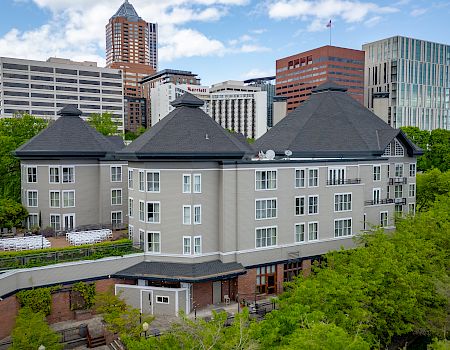A large hotel with a pitched roof sits amid lush trees, with downtown skyscrapers behind, including a Marriott building under a blue sky.