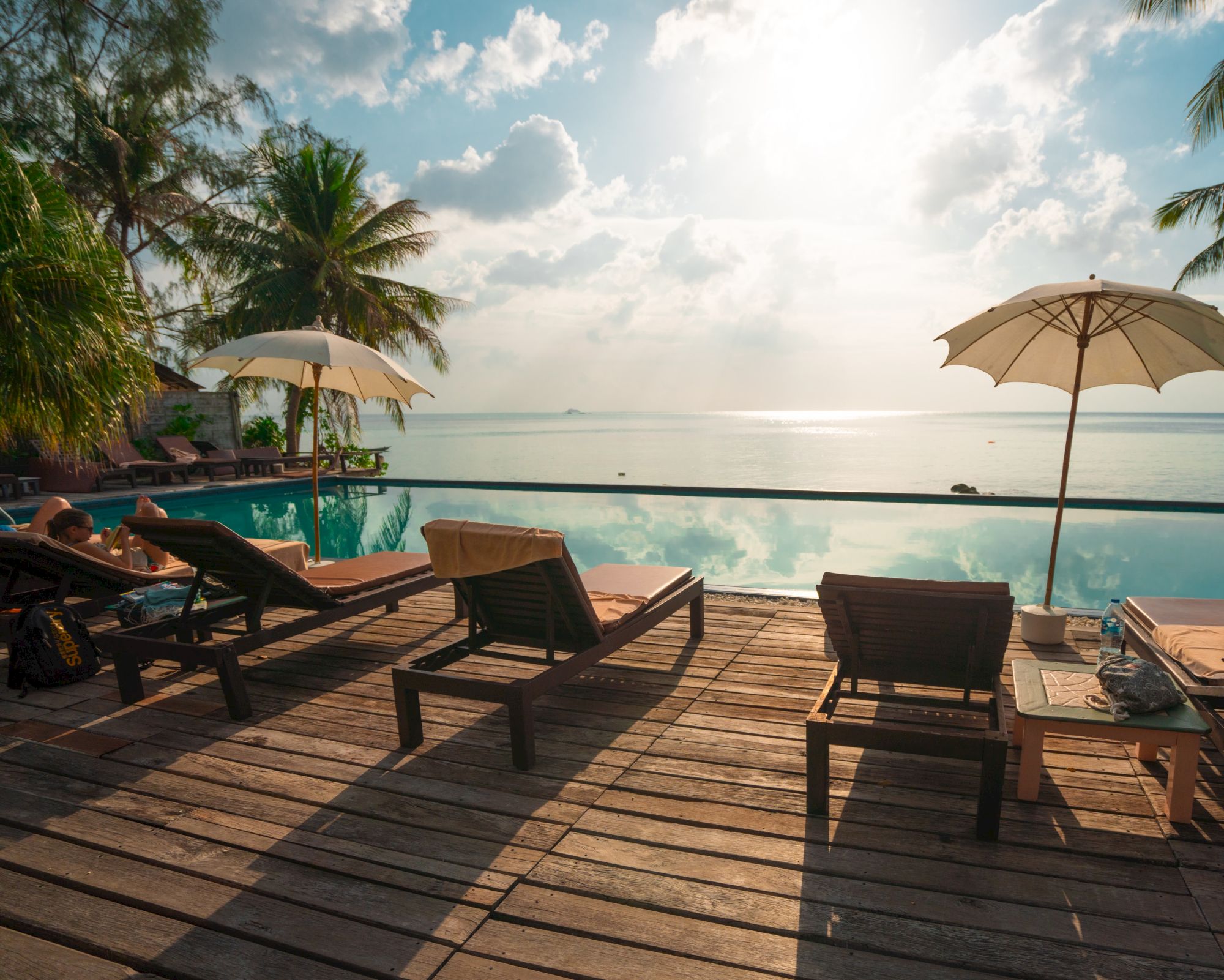 Tropical beachfront resort: a wooden deck with sun loungers, two umbrellas, a pool edge, and palm trees beside the ocean.