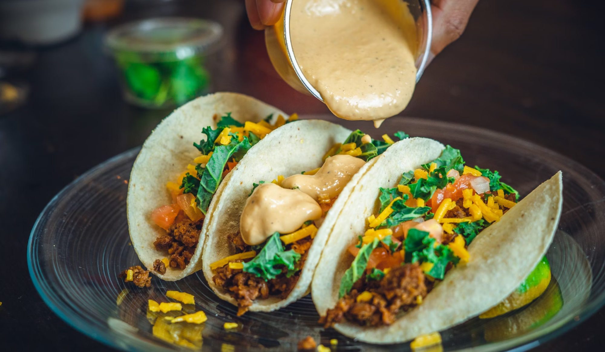 Three tacos on a plate, topped with shredded cheese, diced tomatoes, chopped greens, and a creamy sauce drizzle.