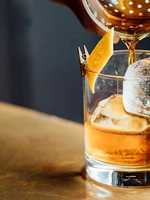 Amber liquor is being poured into a rocks glass over ice, with a lemon peel garnish, on a wooden bar.