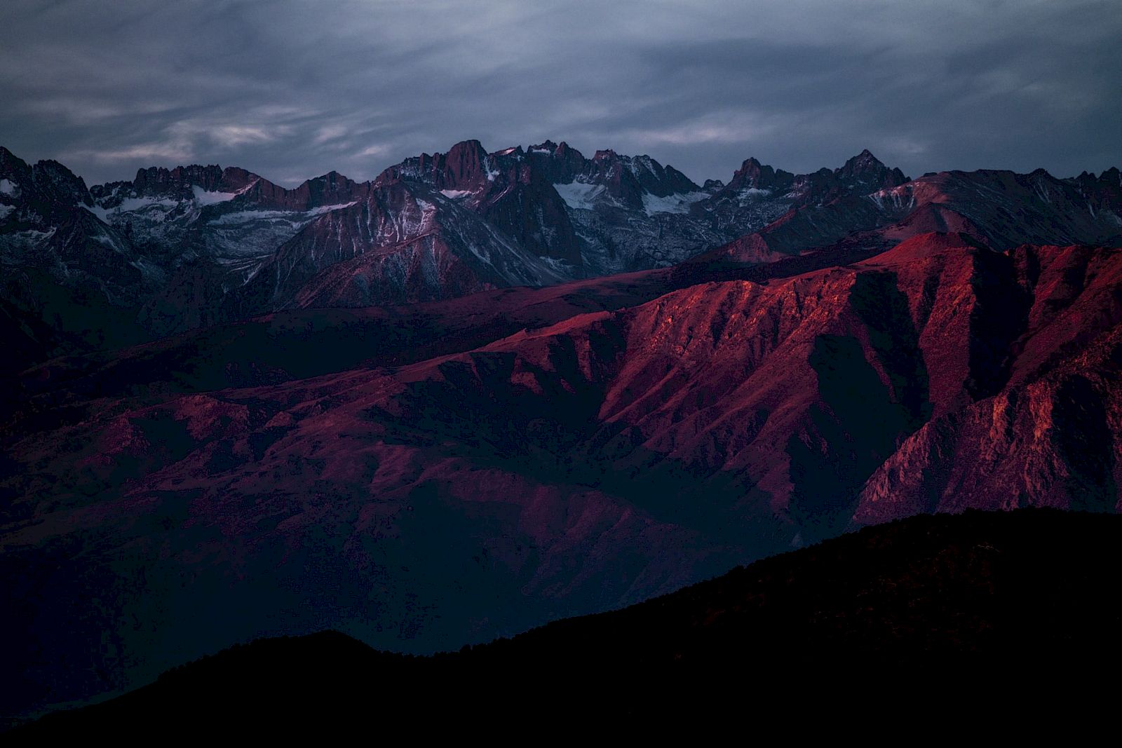 A dramatic mountain range at dusk, with snowy peaks under dark clouds and magenta-lit ridges glowing in the foreground.