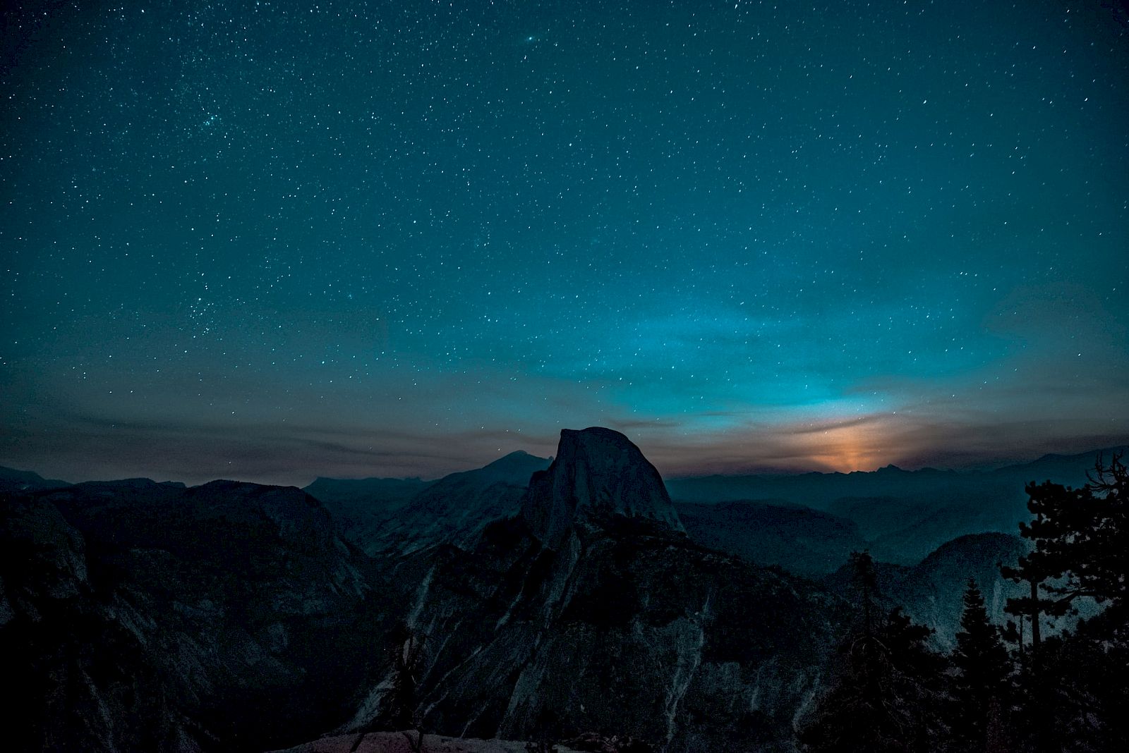 A vast night sky filled with stars over dark mountain peaks, with faint twilight glow near the horizon.