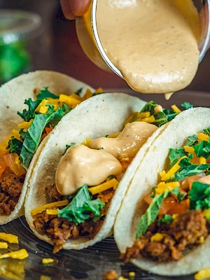 Three tacos on a plate getting drizzled with creamy sauce, topped with chopped cilantro, cheese, and savory meat.