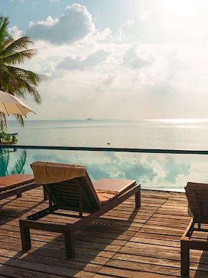 Tropical beachfront view with an infinity pool, lounge chairs, and umbrellas overlooking the ocean and a cloudy sky.