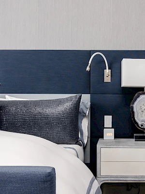 A modern bedroom with a blue tufted bed, pillows, and a nightstand holding a lamp, clock, and remote.