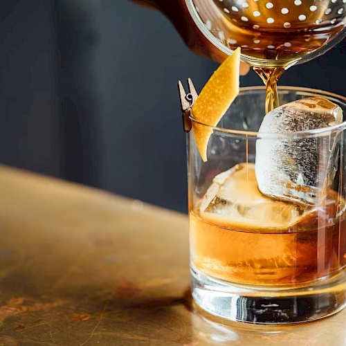 A cocktail being poured into a rocks glass over ice, with amber liquor and a lemon twist garnish. Cheers!