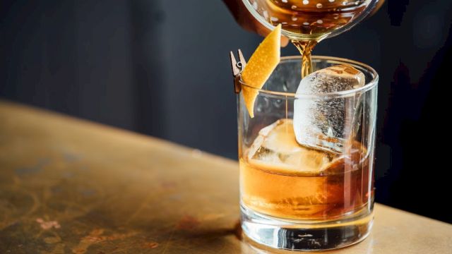 A cocktail being poured into a rocks glass over ice, with amber liquor and a lemon twist garnish. Cheers!