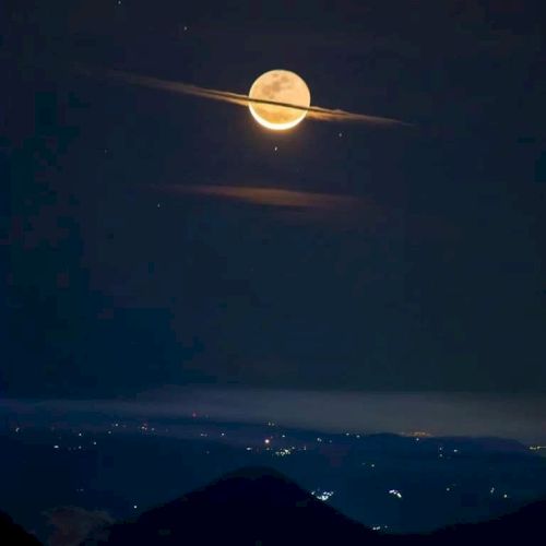 A bright moon glows over dark mountains at night, with stars above and distant city lights far below.
