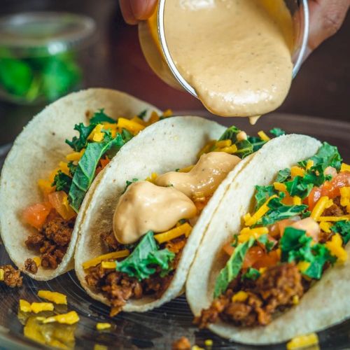 Three tacos on a plate with shredded toppings and sauce being poured over them by hand.