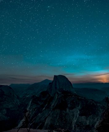 A star-filled night sky over dark mountain silhouettes, with a glowing horizon and a lone peak in the center.