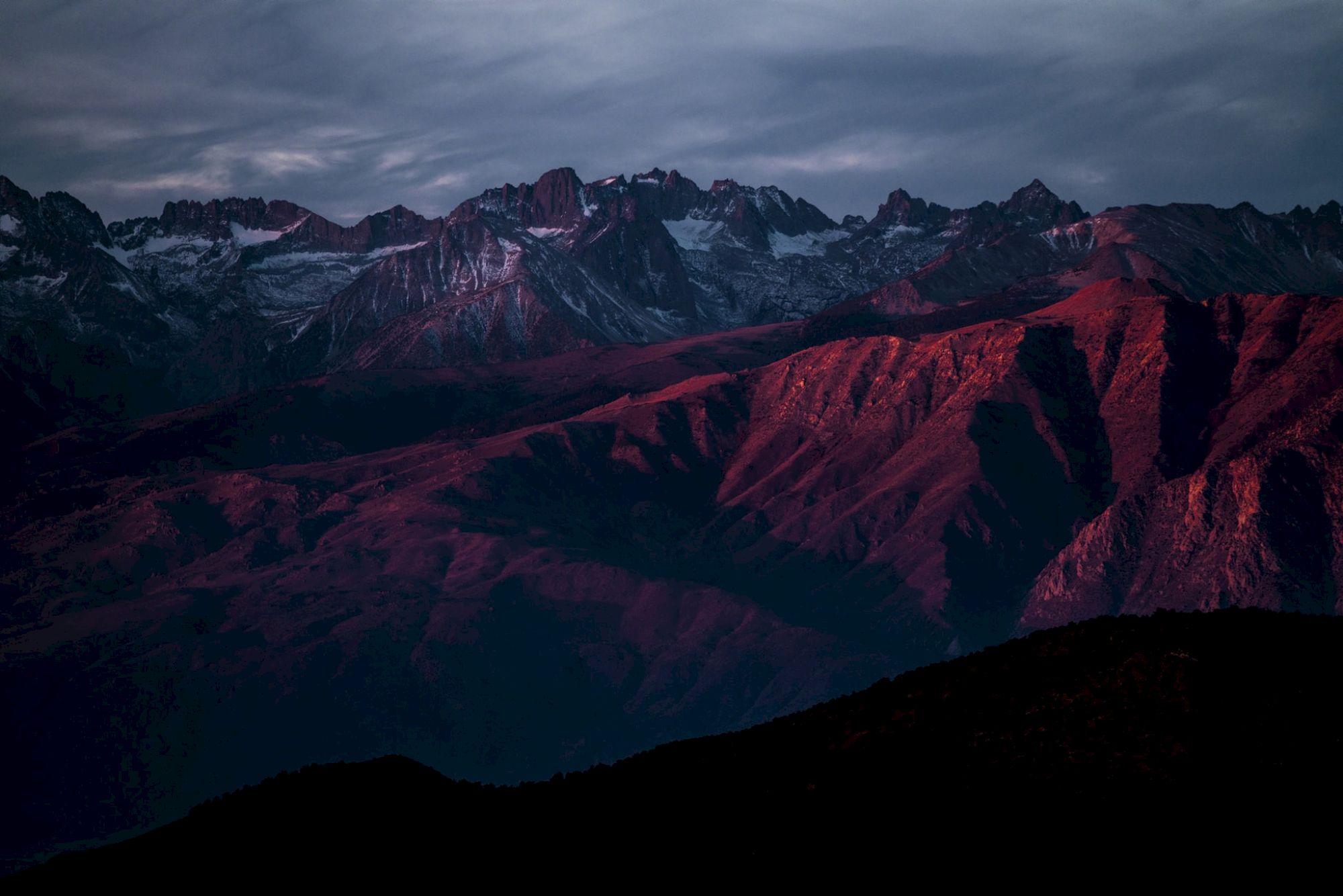Snowy alpine mountain range at dusk, with rugged peaks and a glowing red-orange valley under a cloudy sky.