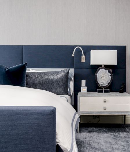 A modern hotel bedroom: blue headboard, pillows, bedside lamp, dresser with decor and a remote on a dark carpet.