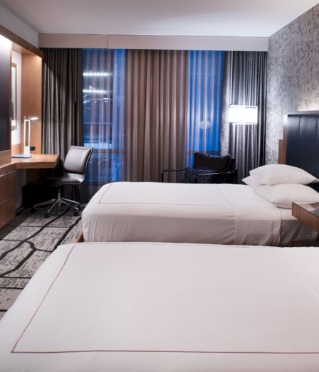 A modern hotel room with two beds, a desk chair, wood panels, and spotlights beside a window with curtains.