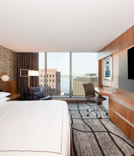 A modern hotel bedroom with a bed, desk chair, built-in TV, wood paneling, and large window view.
