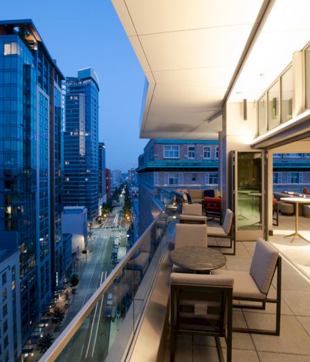 A modern high-rise rooftop terrace with lounge chairs overlooks a city street of office buildings at dusk.