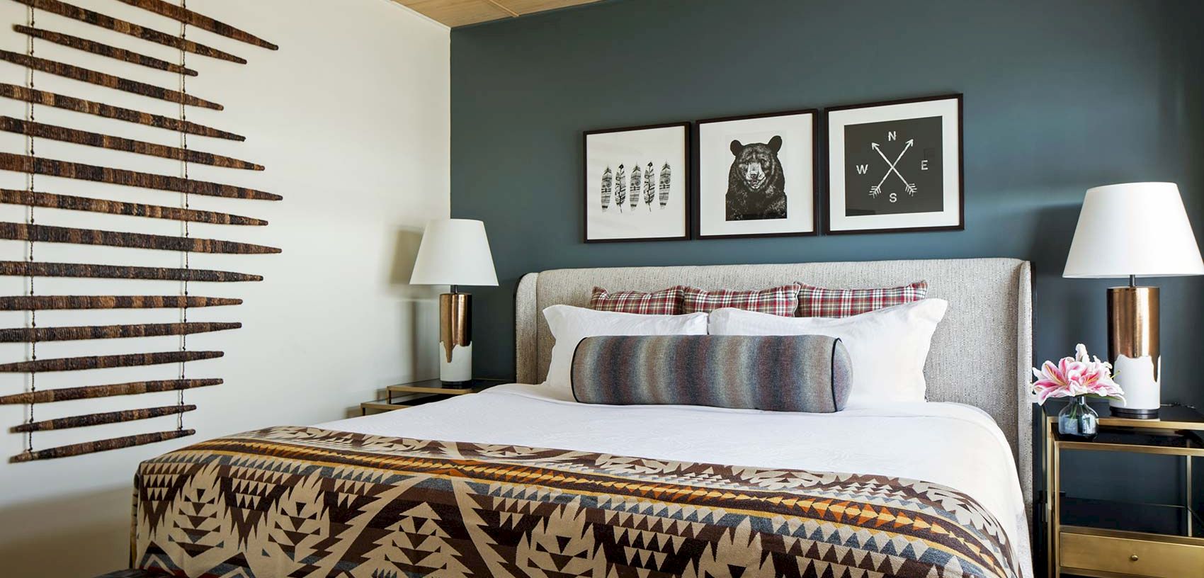 A cozy bedroom with a gray bed, gold lamps, plaid bedding, animal wall art, and wooden blinds.