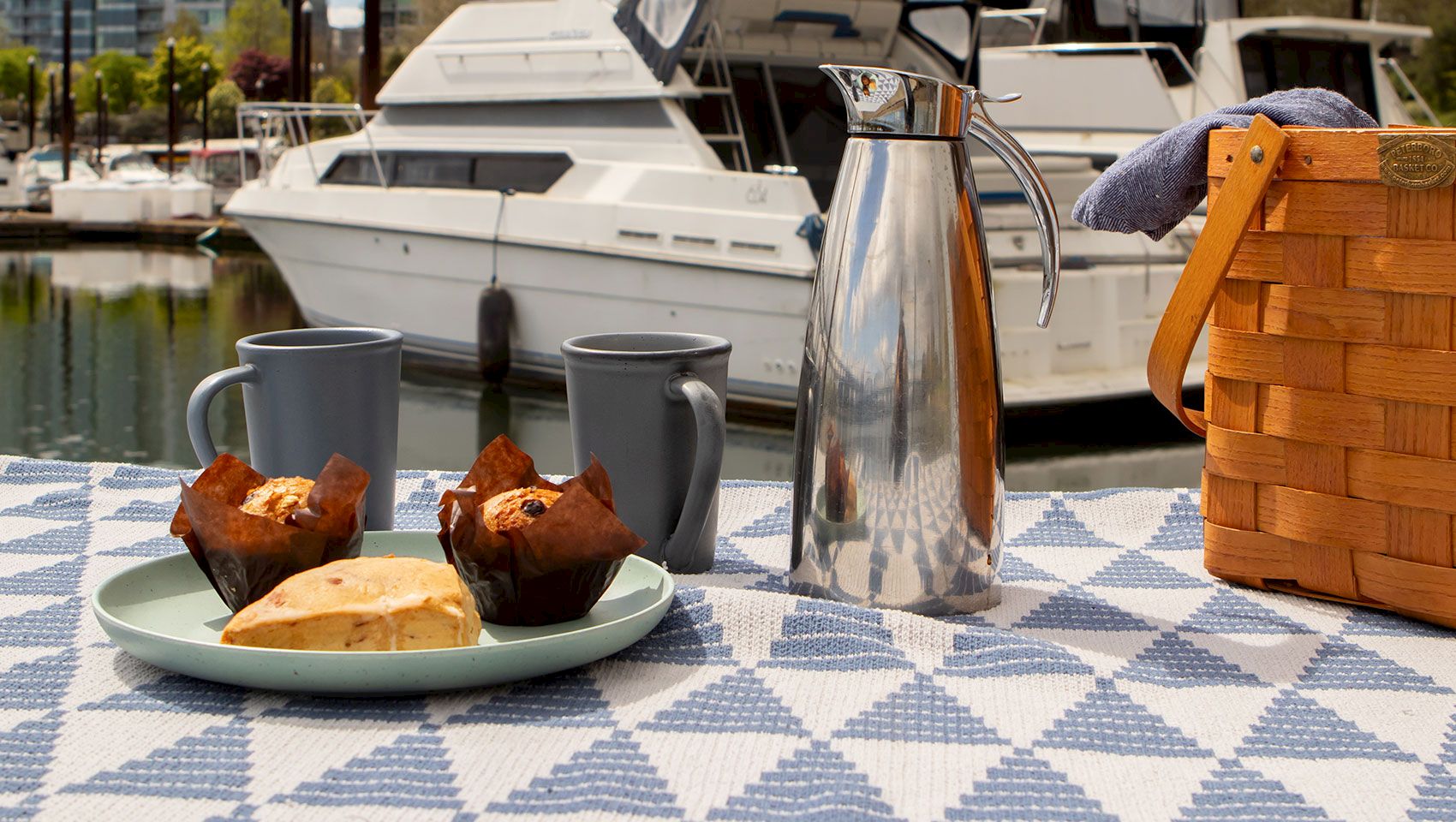 A sunny yacht deck breakfast: silver teapot, two mugs, pastries on a plate, and a wooden basket beside moored boats.