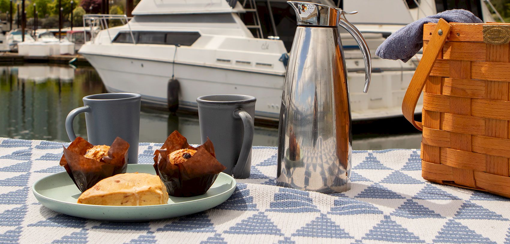 A sunny yacht deck breakfast: silver teapot, two mugs, pastries on a plate, and a wooden basket beside moored boats.