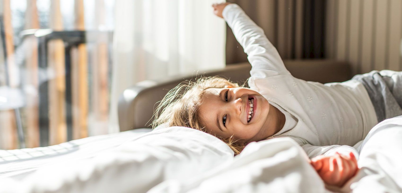 A woman lies in bed smiling, stretching one arm up in a bright, cozy room.