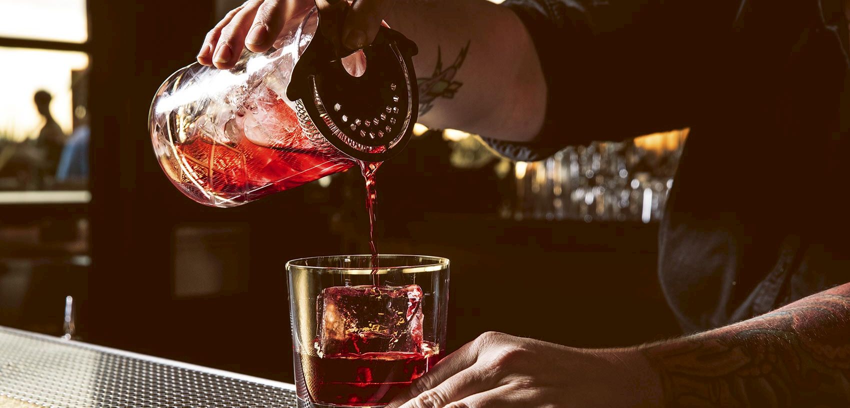 A person pours a red cocktail from a bottle into a tumbler on a patio table at sunset.