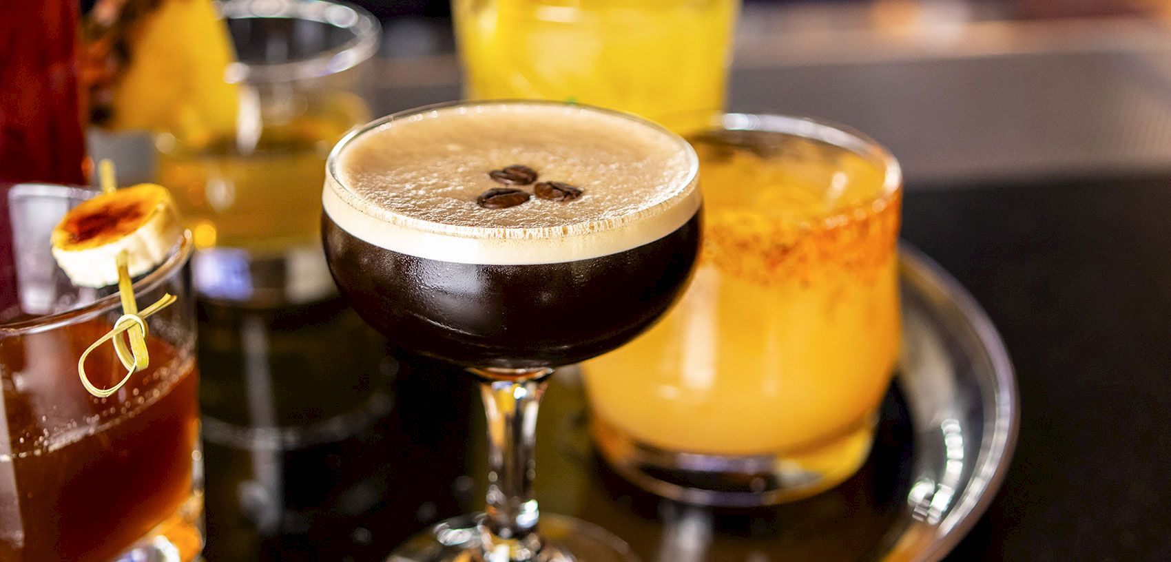 A caf&eacute;-style cocktail in a stemmed glass with coffee beans on top, alongside other amber drinks on a tray.