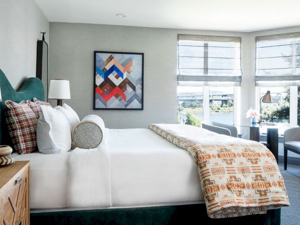 A modern bedroom with a made-up bed, lamps, patterned pillows, and two large windows with blinds.