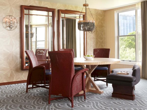 A bright dining room with a round table, red chairs, a beaded chandelier, a wall mirror, and large windows.