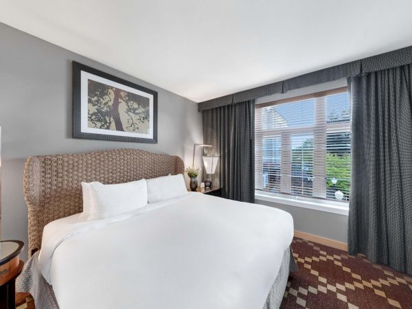 A modern bedroom with a neatly made bed, framed wall art, and dark curtains beside a window with blinds.