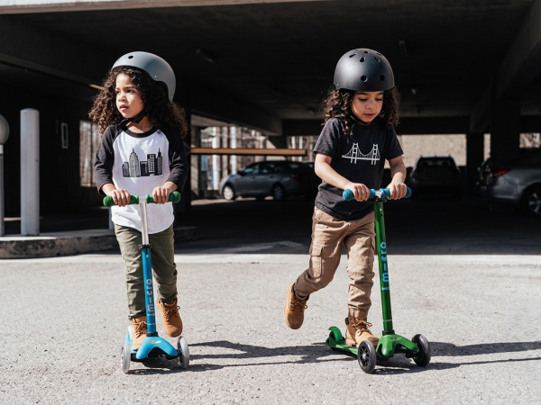 Two kids wearing helmets ride green and blue scooters in a parking garage, one looking forward and the other down.