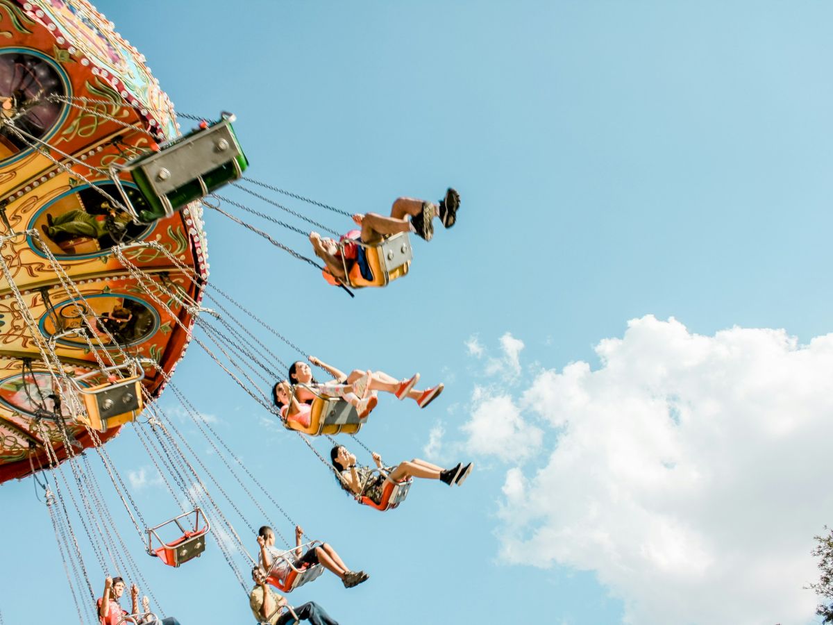 People ride a colorful carnival swing/boat attraction, rising high on long chains above a blue sky.